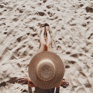 Yaz seyahat moda ve tatil konsepti. Bir hasır şapka ile güzel genç, tabaklanmış kadın yalan ve beyaz kum gösteren bacaklar ile tropikal plajda rahatlatıcı. Üst görünüm, düz yatıyordu.