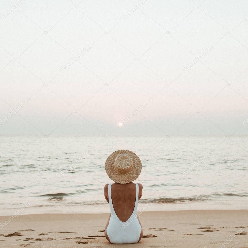 Summer vacation fashion concept. Young, tanned woman wearing a beautiful white swimsuit with a straw hat is sitting and relaxing on tropical beach with white sand and is watching sunset and sea. Vintage or retro tones filter.