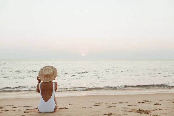 Summer vacation fashion concept. Young, tanned woman wearing a beautiful white swimsuit with a straw hat is sitting and relaxing on tropical beach with white sand and is watching sunset and sea. Vintage warm tones filter.