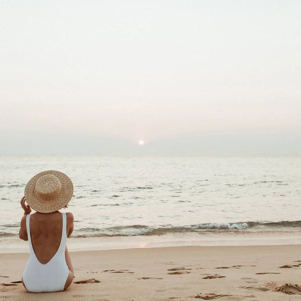 Summer vacation fashion concept. Young, tanned woman wearing a beautiful white swimsuit with a straw hat is sitting and relaxing on tropical beach with white sand and is watching sunset and sea. Vintage warm tones filter.