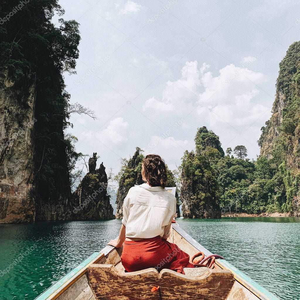 Young woman in red skirt and white blouse sitting on wooden boat watching at exotic and tropical dark green big islands with rocks and turquoise lake at Cheow Lan Lake, Khao Phang, Ban Ta Khun District, Thailand. Travel holiday and adventure concept.