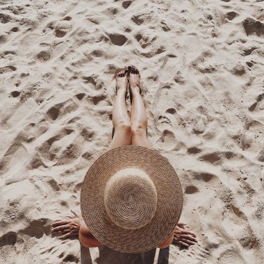 Yaz seyahat moda ve tatil konsepti. Bir hasır şapka ile güzel genç, tabaklanmış kadın yalan ve beyaz kum gösteren bacaklar ile tropikal plajda rahatlatıcı. Üst görünüm, düz yatıyordu.