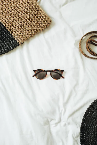 Women\'s fashion accessories with glasses in the middle of the photo and belt, straw hat, straw bag lying on bed with white sheet. Summer and vacation concept. Top view, flat lay.