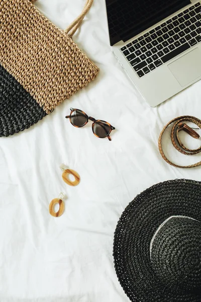 Women\'s fashion accessories: glasses, earrings, belt, straw hat, straw bag and laptop lying on bed with white sheet. Summer and travel concept. Top view, flat lay.