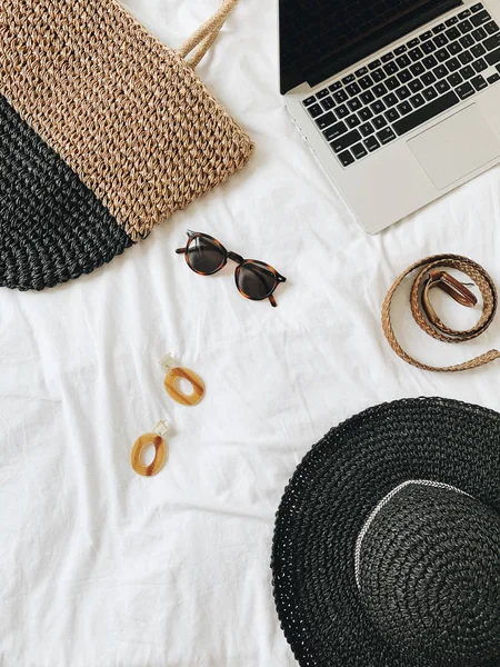 Women\'s fashion accessories: glasses, earrings, belt, straw hat, straw bag and laptop lying on bed with white sheet. Summer and travel concept. Top view, flat lay.