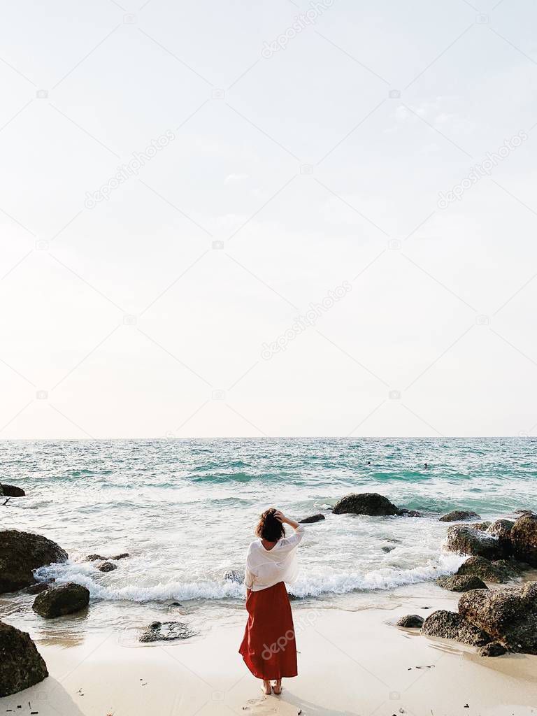 A beautiful young woman in red skirt and white shirt standing on the beach watching at blue sea with waves, rocks and cream sky. Open space for text. Summer and travel concept.