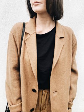 Sonbahar, sonbahar ve moda konsepti. Kahverengi saçlı, bej paltolu, siyah gömlekli, beyaz duvara karşı duran kahverengi pantolonlu genç bir kadın. Nötr renklere sahip yakın çekim fotoğraf.