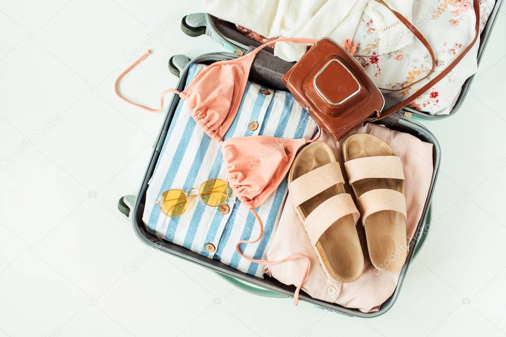 Flat lay of luggage with summer clothes and accessories: bikini, slippers, retro camera, sunglasses, dress on white background. Top view fashion travel composition.