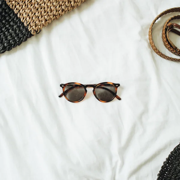 Women\'s fashion accessories with glasses in the middle of the photo and belt, straw hat, straw bag lying on bed with white sheet. Summer and travel concept. Top view, flat lay.