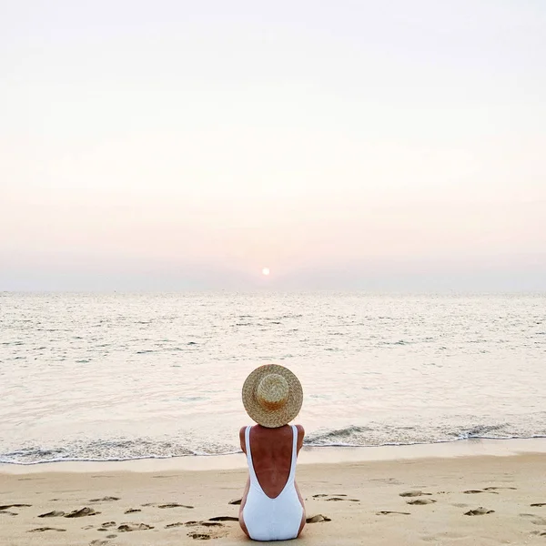 Summer holiday fashion concept. Young, tanned woman wearing a beautiful white swimsuit with a straw hat is sitting and relaxing on tropical beach with white sand and  watching colorful pink sunset and sea. Vintage or retro tones filter.