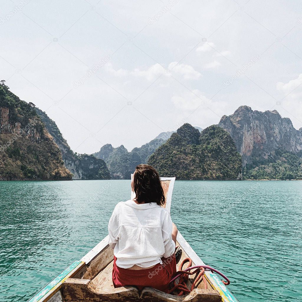 Young woman in red skirt and white blouse sitting on wooden boat watching at exotic and tropical dark green big islands with rocks and turquoise lake at Cheow Lan Lake, Khao Phang, Ban Ta Khun District, Thailand. Travel holiday and adventure concept.