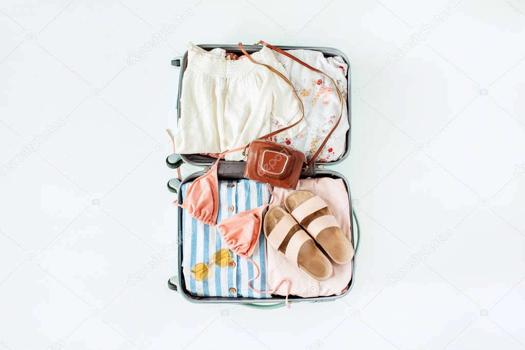 Hand luggage with bikini, sunglasses, slippers, retro camera and dress on white background. Flat lay, top view travel vacation fashion composition.