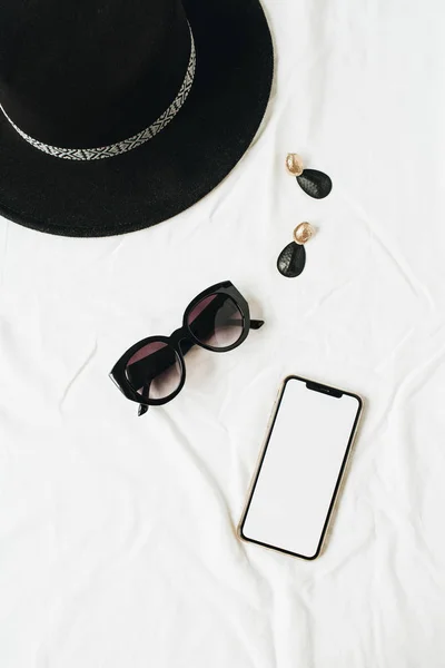 Fashion mock up background with women's accessories on white linen. Hat, sunglasses, earrings, smart phone with blank copy space screen. Flat lay, top view minimal lifestyle fashion blog template.