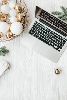 Noel / Yeni Yıl / Noel kompozisyonu. Düz yerde, üst tarafta. Laptop, Noel ağacı dallarıyla süslenmiş beyaz battaniyenin üzerinde duruyor. Beyaz ve altın toplardan oluşan bir sepet, kış şapkası ve beyaz eldivenler. Ticari serbest konsept.