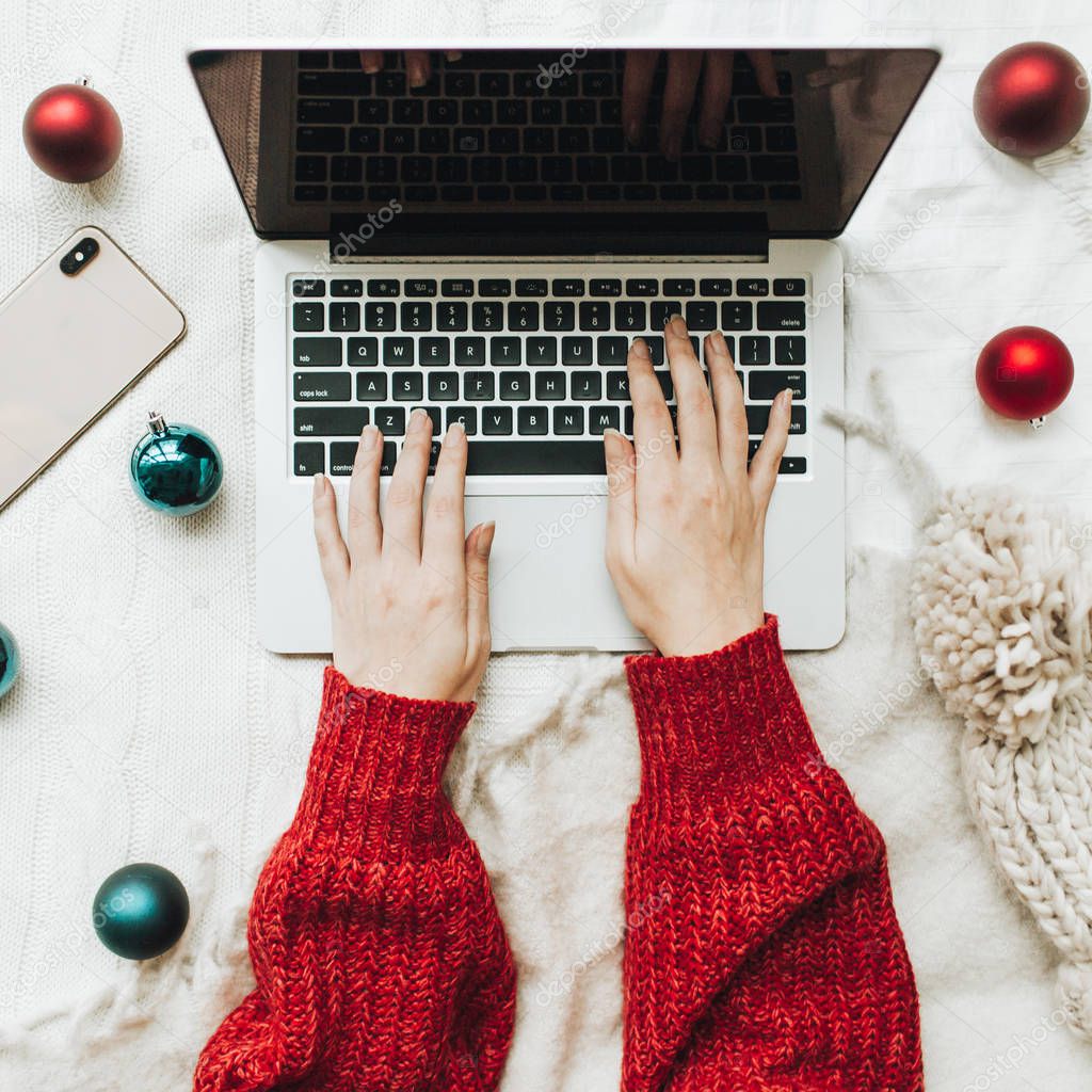 Christmas / New Year composition. Flat lay, top view. Young woman hands in red knitted sweater typing on laptop on the white bed with white blanket decorated with christmas red and blue balls and winter knitted hat. Minimal business freelance concept