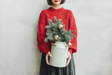 Kırmızı örülmüş süveteri ve elinde gri eteği olan genç ve güzel bir kadın. Altın toplarla süslenmiş noel ağacından yapılma çam ağacından yapılmış sürahiyi ellerinde tutuyor. Minimum stil noel ve yeni yıl konsepti.