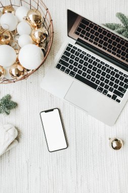 Noel / Yeni Yıl / Noel kompozisyonu. Düz konum, üst görünüm, maket, kopyalama alanı. Noel ağacı dallarıyla süslenmiş beyaz battaniyenin üzerinde duran cep telefonlu dizüstü bilgisayar, beyaz ve altın toplarla dolu bayram sepeti. Ticari serbest konsept.