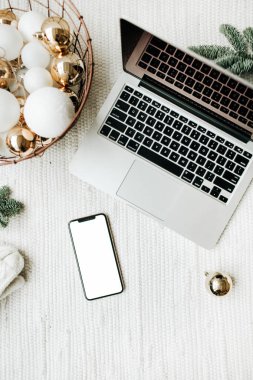 Noel / Yeni Yıl / Noel kompozisyonu. Düz konum, üst görünüm, maket, kopyalama alanı. Noel ağacı dallarıyla süslenmiş beyaz battaniyenin üzerinde duran cep telefonlu dizüstü bilgisayar, beyaz ve altın toplarla dolu bayram sepeti. Ticari serbest konsept.