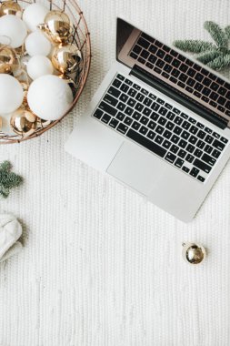 Noel / Yeni Yıl / Noel kompozisyonu. Düz yerde, üst tarafta. Laptop, Noel ağacı dallarıyla süslenmiş beyaz battaniyenin üzerinde duruyor. Beyaz ve altın toplardan oluşan bir sepet, kış şapkası ve beyaz eldivenler. Ticari serbest konsept.