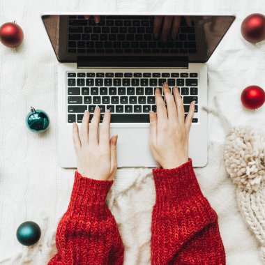 Noel / Yeni Yıl kompozisyonu. Düz yerde, üst tarafta. Genç kadın elleri kırmızı örülmüş süveter içinde beyaz yatakta dizüstü bilgisayarda yazıyor. Kırmızı ve mavi toplar ve kışın örülmüş şapkayla süslenmiş beyaz battaniyeyle. Asgari iş serbest konsepti
