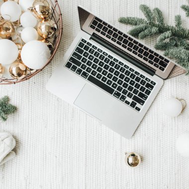 Noel / Yeni Yıl / Noel kompozisyonu. Düz yerde, üst tarafta. Laptop, Noel ağacı dallarıyla süslenmiş beyaz battaniyenin üzerinde duruyor. Beyaz ve altın toplardan oluşan bir sepet, kış şapkası ve beyaz eldivenler. Ticari serbest konsept.