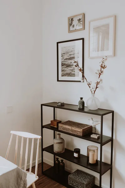 Modern scandinavian nordic living room with beautiful details such as black shelves, vases, frames, wooden caskets, cotton, wooden beige chair, white table. Scandi design interior concept.