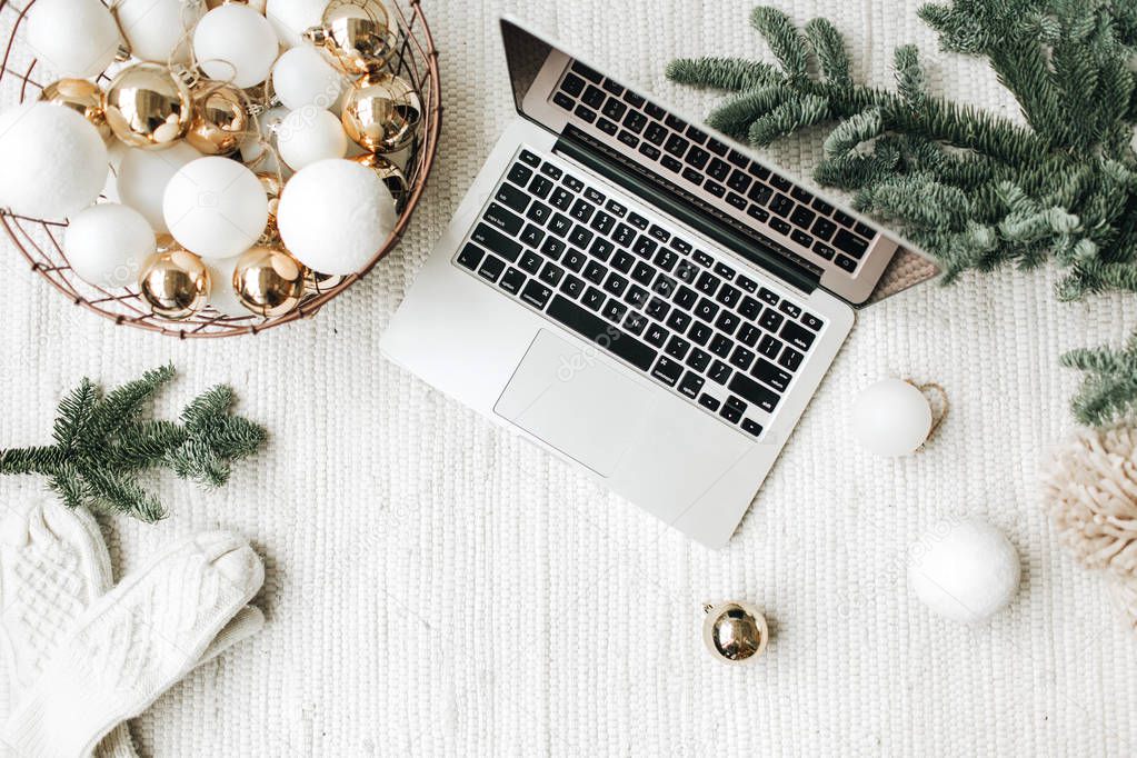 Christmas / New Year / xmas composition. Flat lay, top view. Laptop lying on the white blanket decorated with christmas tree branches, festive basket of white and gold balls, winter hat and white mittens. Business freelance concept.