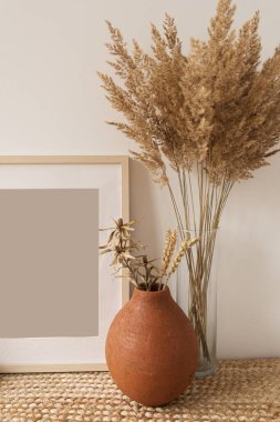 Blank photo frame with copy space, reeds bouquet in vase, rye in clay pot at white wall. Mockup template. Modern interior design concept. clipart