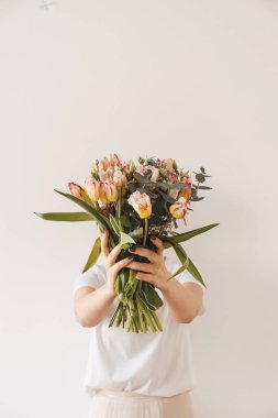 Beyaz bluzlu genç ve güzel bir kadın lale çiçeği buketini elinde beyaz duvara karşı tutuyor. Bayram kutlaması çiçek konsepti