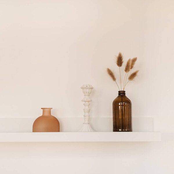 White shelf on white wall with bottle, vase, fluffy pom pom plants, candlestick. Minimal modern interior design concept.