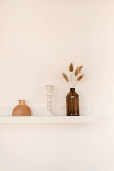 White shelf on white wall with bottle, vase, fluffy pom pom plants, candlestick. Minimal modern interior design concept.