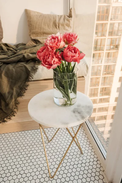 Beautiful pink peony flowers bouquet in glass vase on marble table. Modern stylish interior design concept with wooden lounger, mosaic tiles, pillows, plaid