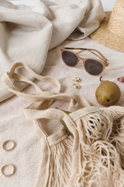 Neutral fashion composition with women's accessories and bijouterie on beige blanket. String bag, straw hat, sunglasses, rings, earrings, pear. Minimal lifestyle concept.