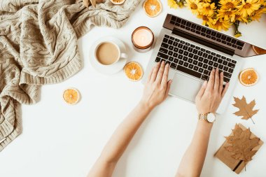 Kadın dizüstü bilgisayarda çalışıyor. Ofis masası çalışma alanı. Düz yatış, üst düzey bayan patron iş konsepti. Klavyede kadın eli var. Battaniye, çiçek buketi, yapraklar, beyaz arka planda kahve fincanı.