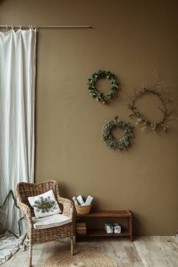 Zeytin duvarındaki köknar iğneleri ve dallardan yapılmış yuvarlak ev yapımı çelenk çerçeveleri. Noel kutlaması süsü. Yeni yıl kompozisyonu. Rattan sandalyeli ve mobilyalı modern iç tasarım..