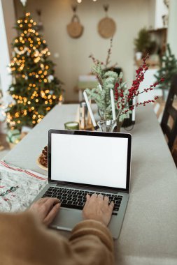 Genç bir kadın dizüstü bilgisayarda çalışıyor. Boş ekran ve taklit alanı var. Konforlu mutfak, masa, Noel ağacı, köknar dalları, koniler, mumlar. Tatil kutlamaları.