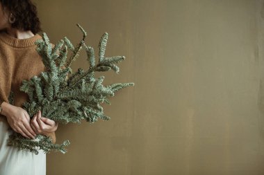 Süveter ve etekli genç bir kadın zeytin duvarına köknar dalları tutuyor. Minimalist Noel / Yeni Yıl konsepti.