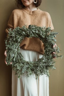 Süveter ve etekli genç bir kadın zeytin duvarına karşı köknar dallarından yapılmış yuvarlak bir çelenk tutuyor. Minimum moda festivali Noel / Yeni Yıl Kutlama Konsepti. 