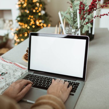 Genç bir kadın dizüstü bilgisayarda çalışıyor. Boş ekran ve taklit alanı var. Konforlu mutfak, masa, Noel ağacı, köknar dalları, koniler, mumlar. Tatil kutlamaları.
