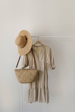 Hasır şapka, saman el çantası, beyaz duvardaki elbise askılığında bej keten elbise. Moda giysileri, online alışveriş konsepti.