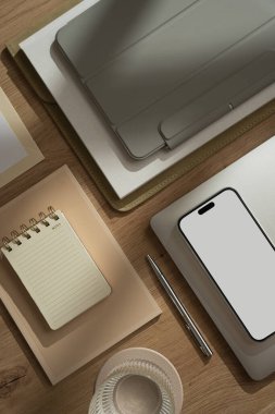 Boş ekranlı cep telefonu, dizüstü bilgisayarı, tablet defteri, bir bardak su, defter, belgeler, güneş ışığı gölgeli ahşap masa üzerinde fotokopi alanı olan boş kağıt sayfası olan estetik ofis çalışma alanı.