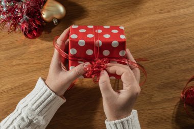 Beyaz süveter içinde kadın elleri kırmızı benekli kağıt ve kurdele ile Noel hediyesini ahşap masa, bayram hazırlığı, üst manzara