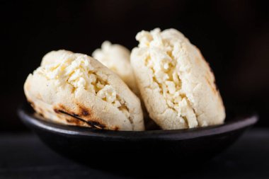 Kolombiyalı geleneksel beyaz Mısır arepa siyah arka plan üzerine siyah bir seramik tabak içinde rendelenmiş peynir ile doldurulmuş