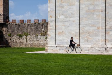 Woman biking next to the ancient walls of Pisa and Monumental Cemetery at the Square of Miracles in a beautiful early spring day clipart