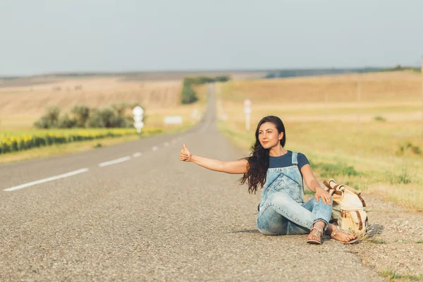 hipster girl with  backpack sitting on  side of  road. concept - hitchhiking, adventure, freedom. Copy space