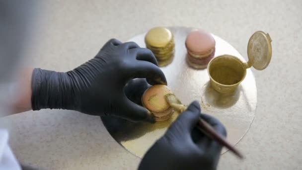 Mains de femme en gants noirs décorant macaron rose avec éclaboussure d'or en utilisant une brosse dans la cuisine — Video