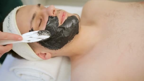 メンズ顔のためのブラックマスク。スパで黒いマスクを及ぼしによって適用されるリラックスした若い男のクローズアップビュー. — ストック動画