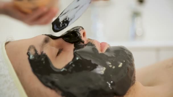 L’esthéticienne met un masque noir sur l’homme, en appliquant un masque cosmétique rajeunissant de tissu noir au visage — Video