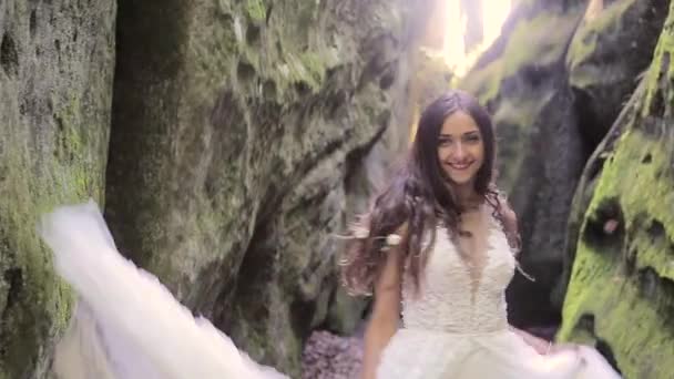 Linda novia en un vestido de novia corre entre las rocas, y mira a la cámara con una gran sonrisa. Rostros felices, emociones — Vídeo de stock