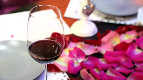 Ένα ποτήρι κόκκινο κρασί στέκεται πάνω στο τραπέζι που καλύπτεται με πέταλα από τριαντάφυλλα — Αρχείο Βίντεο